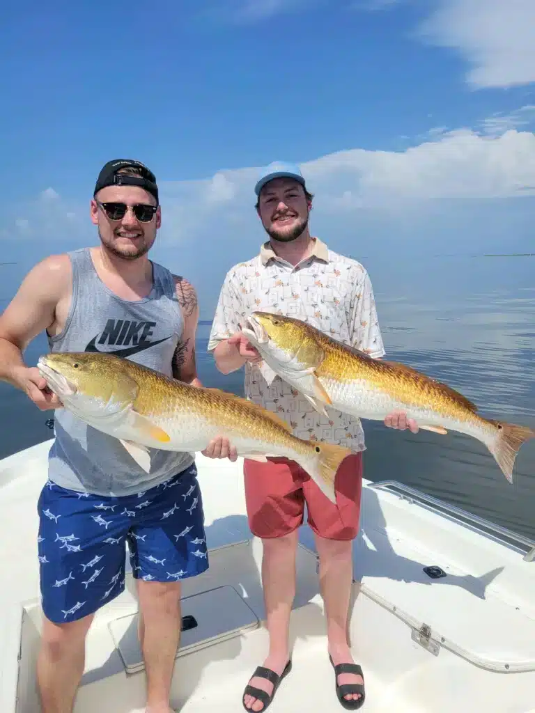 Two men on a fishing boat in the bayou, each holding redfish they caught on a New Orleans Style Fishing Charter fishing trip